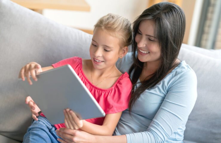 Mom and daughter relaxing on the couch looking at videos on a digital tablet both smiling